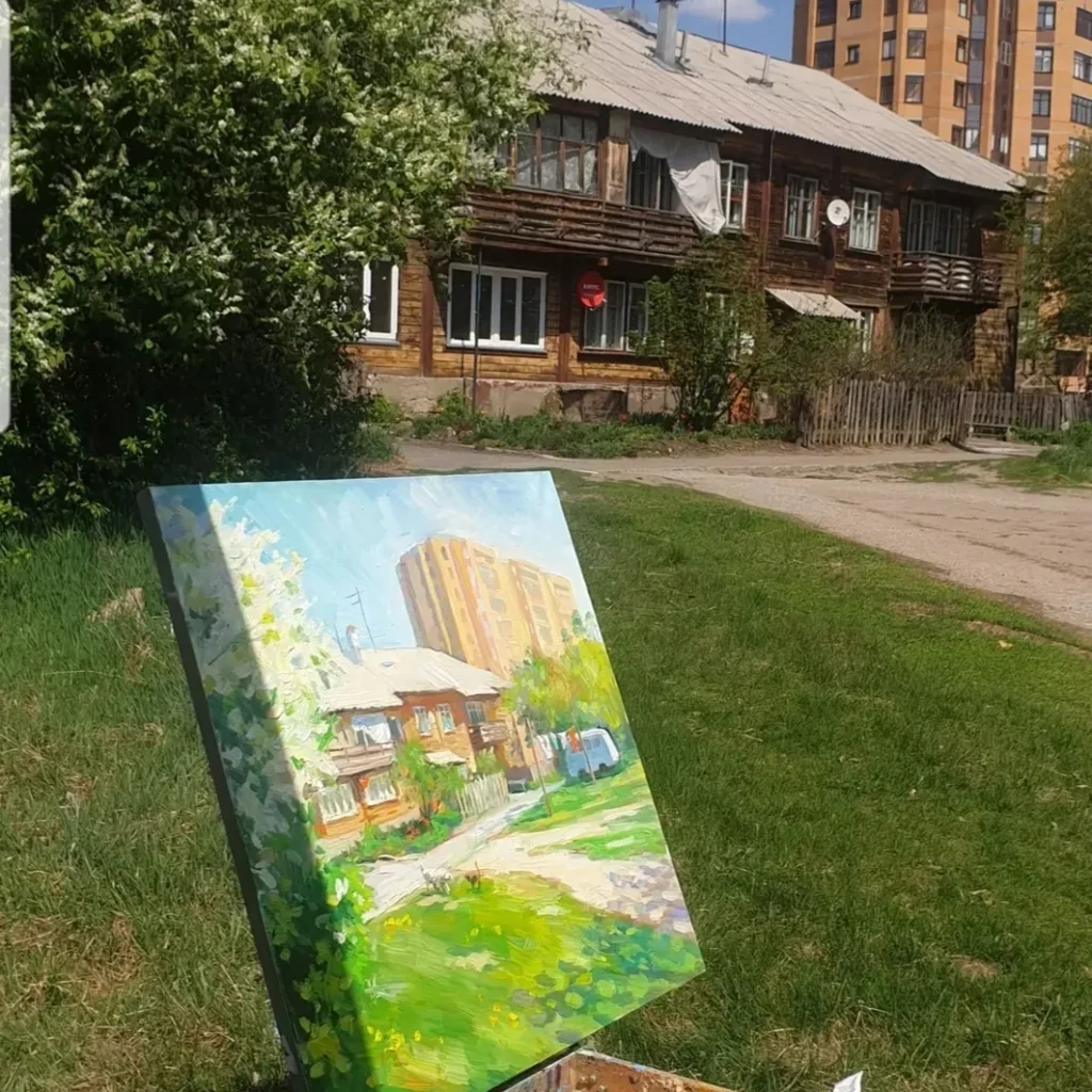 Картина «Среди небоскребов», установленная на этюднике на фоне реального пейзажа. На холсте запечатлен момент завершения работы: яркая палитра свежих мазков передает сочную зелень и свет весеннего дня, точно повторяя очертания старого деревянного дома и возвышающейся за ним многоэтажки. Удивительное сходство живописи и натуры подчеркивает искренность момента и ценность уходящей истории этого дворика.