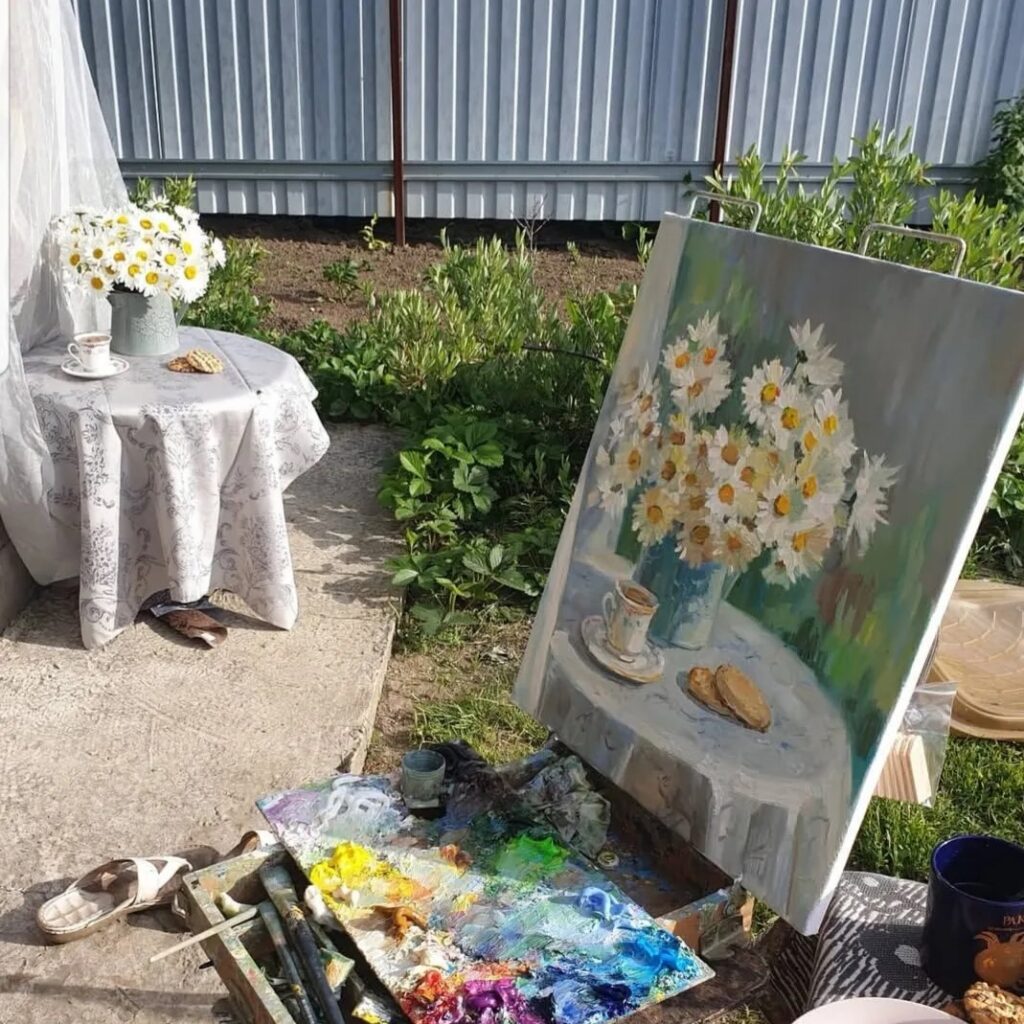 Картина «Чай с ромашкой» на этюднике на фоне реального натюрморта, послужившего натурой: круглого стола с букетом ромашек и чашкой чая в саду. На переднем плане видна деревянная палитра, густо покрытая яркими масляными красками — ультрамарином, кадмием желтым и белилами. Холст точно передает мягкое утреннее освещение и игру теней на скатерти, создавая полное единство написанного изображения и живого момента в саду.